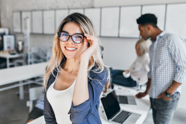 Indoor portrait of excited student girl in glasses and gray jacket. Attractive female employee posing in office and laughing with colleagues on background.