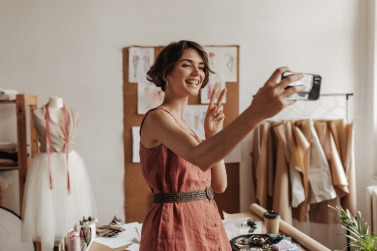 pretty-young-brunette-short-haired-woman-linen-red-dress-with-black-belt-smiles-takes-selfie-shows-peace-sign-poses-office-fashion-designer